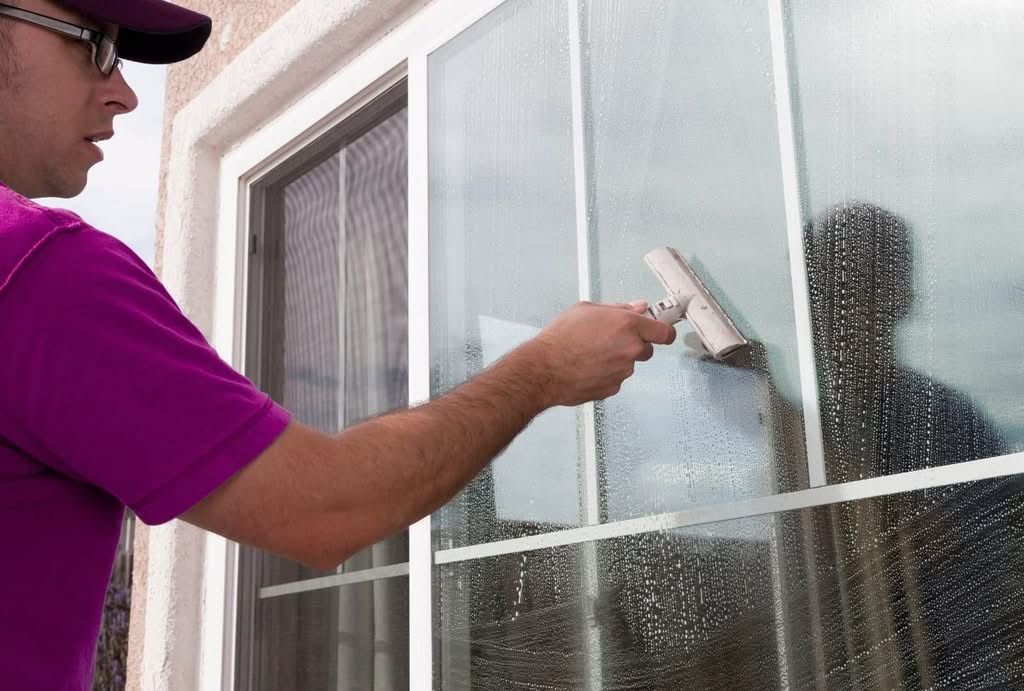 A close-up of a worker in a purple/pink shirt, baseball cap, and glasses, using a blue and white fabric scrubber to apply soapy water to a glass window with white trim. The background is a sunny yard with plants. Commercial window cleaning in Florida.