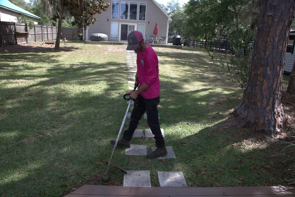 A man in a bright pink long-sleeve shirt, dark pants, and a baseball cap is operating a stand-on lawnmower in a grassy area next to a paved road. He is looking back over his shoulder towards the camera, with a white car and a trailer visible on the road behind him.
