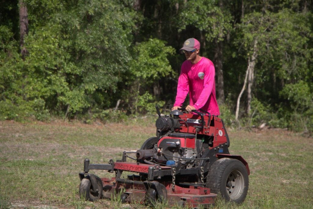 A man in a pink long-sleeve shirt, jeans, and a baseball cap is using a string trimmer (weed wacker) to trim grass along a pathway of concrete stepping stones in a sunny backyard. A large house and trees are visible in the background. Doing commercial lawn care in Silver Springs Shores, Florida and OTOW Lawn Care.