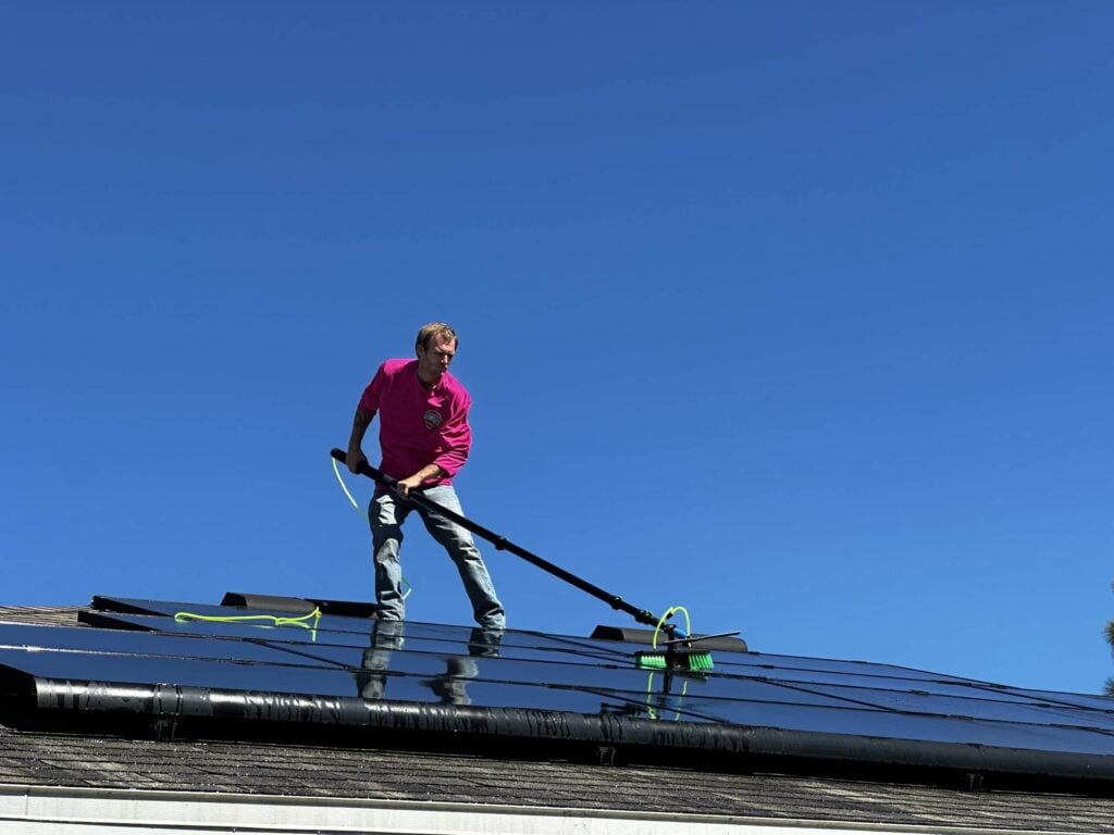 A man in a pink shirt and jeans is standing on a dark, reflective rooftop covered in solar panels, using a long pole with a brush attachment to clean them under a bright blue sky.