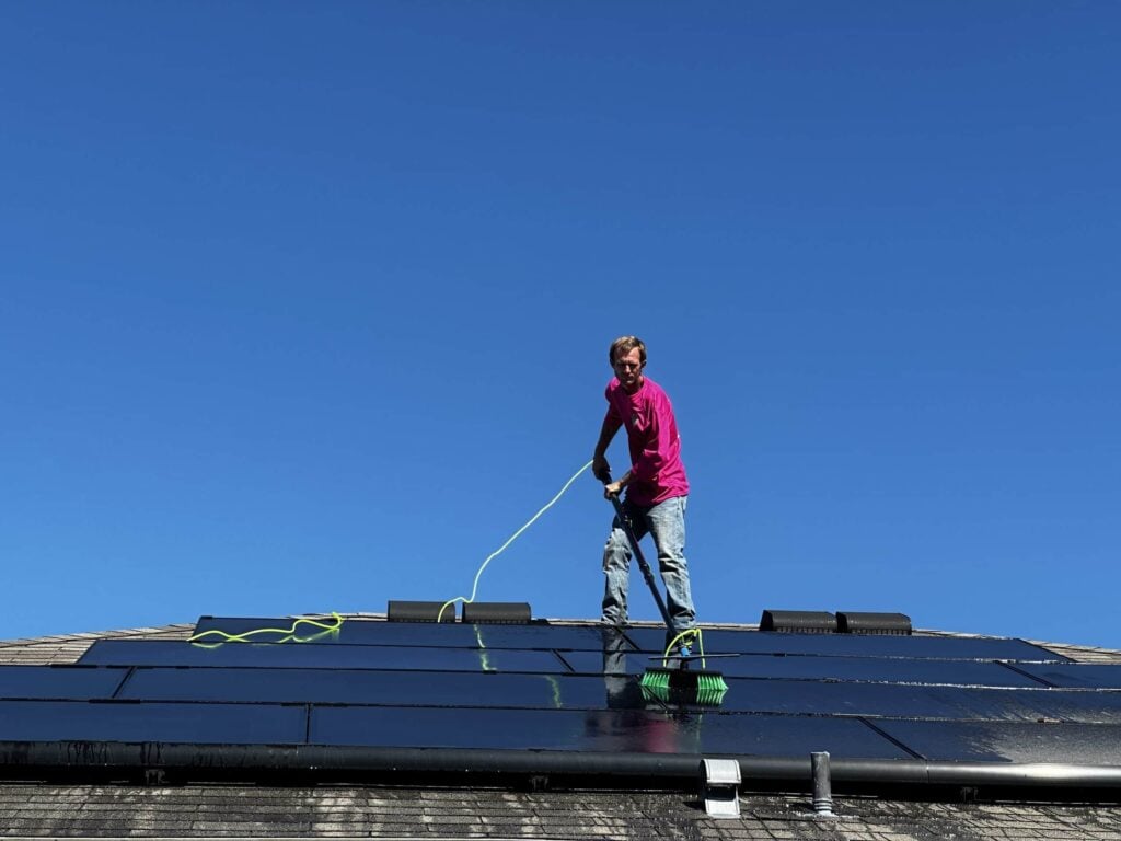 Professional Solar panel cleaning. A man's hand, wearing a pink long-sleeve shirt, reaches down towards the pavement to pick up a small, crumpled piece of trash or residue (possibly chewed gum). The background shows a blurry street and buildings at sunset.