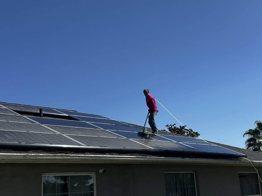 A worker in a pink long-sleeve shirt is using a long brush on a pole, connected to a hose, to clean dark, reflective solar panels installed on a residential roof under a bright blue sky. The background shows the house's siding and window.