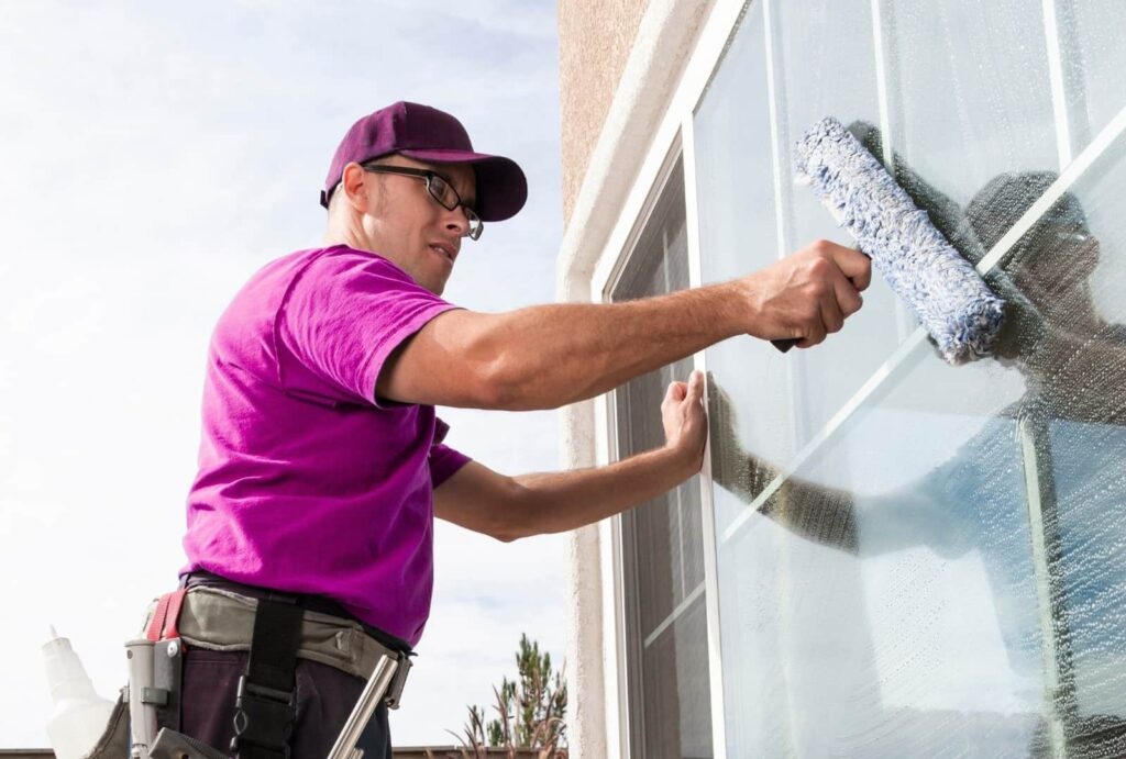 A worker in a pink t-shirt, dark cap, and glasses is pouring cleaning solution from a white bottle onto a window cleaning tool with a blue and white fabric head. He is doing window cleaning a large glass window of a house on a sunny day.