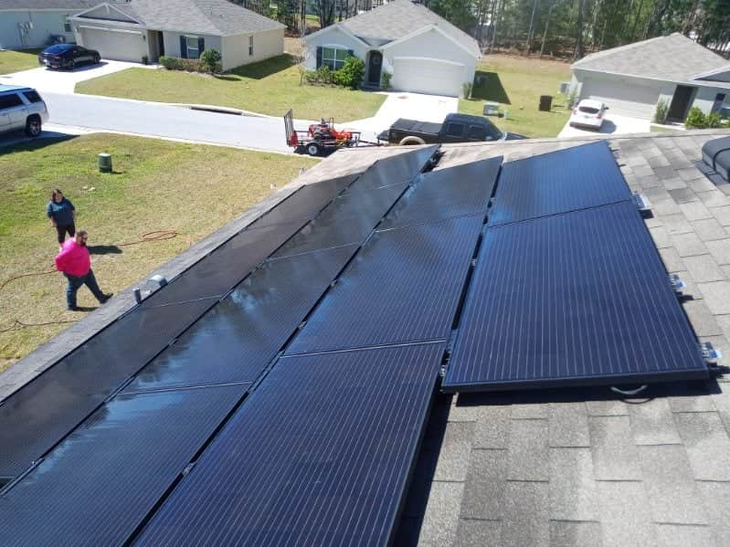 An aerial view of a residential roof covered in dark shingles with a large array of reflective black residential solar panel cleaning. Two workers, one in a pink shirt, are visible on the left edge of the roof. The background shows suburban houses, driveways, and a trailer attached to a truck.
