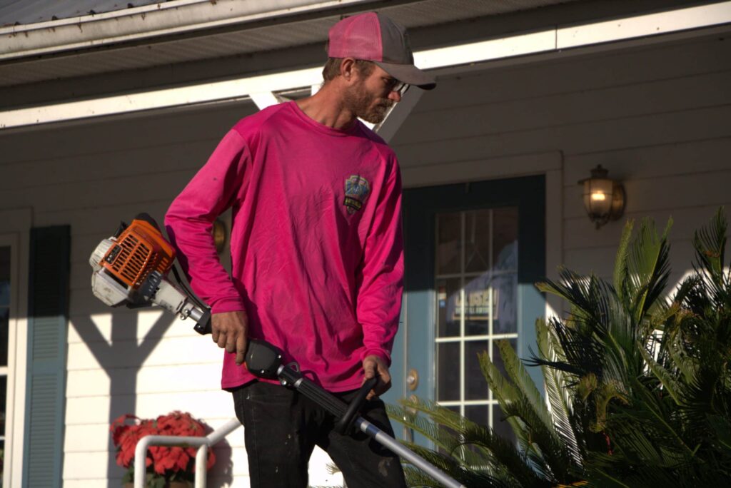A professional landscaper in a pink long-sleeve shirt and grey baseball cap using a gas-powered string trimmer to edge a residential yard near a house.