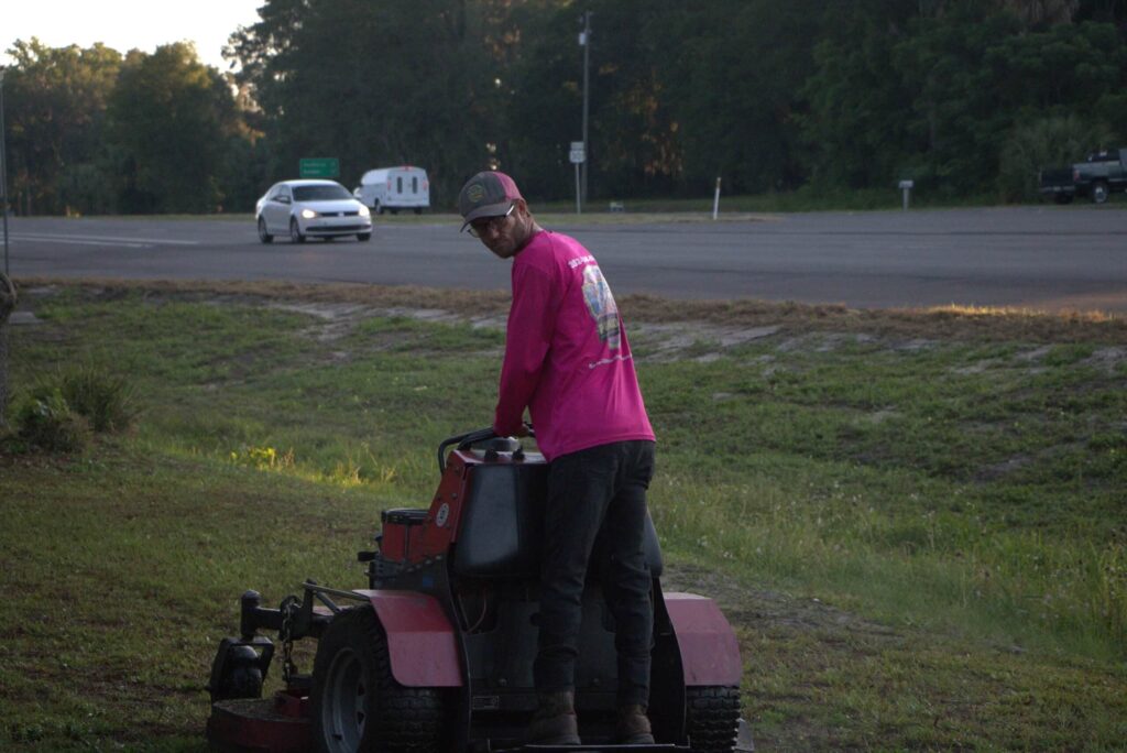 A professional landscaper in a high-visibility pink shirt operating a red stand-on lawnmower on a grassy shoulder next to a paved highway.
