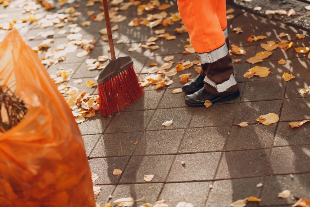 A street cleaner in high-visibility orange trousers, professional parking lot sweeping and reflective boots sweeping dry autumn leaves on a paved sidewalk with a red broom.