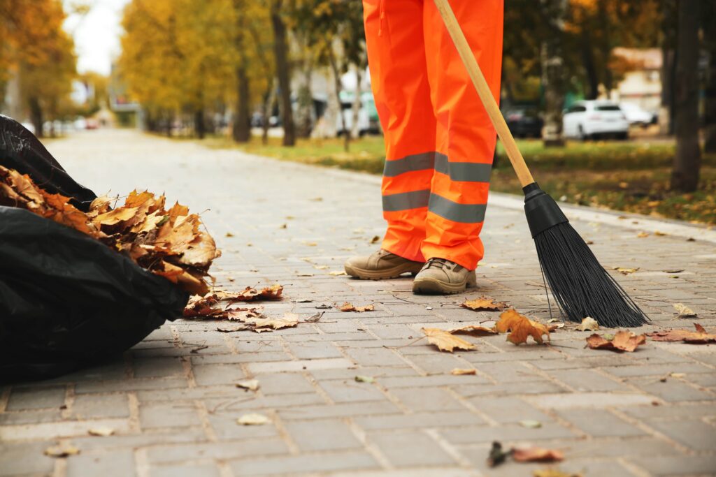 A street cleaner in bright orange high-visibility safety gear using a large broom to commercial lot sweeping fallen autumn leaves into a black waste bag on a paved park walkway.