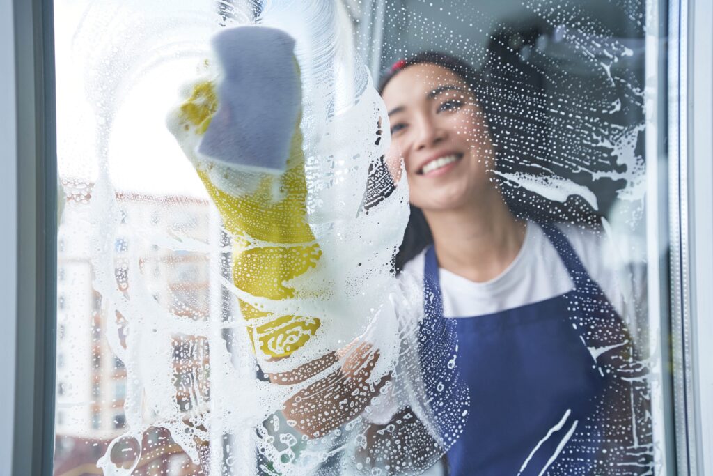 A smiling young woman wearing yellow rubber gloves and a blue apron uses a sponge to scrub soapy lather onto a glass professional window cleaning panel from the outside.