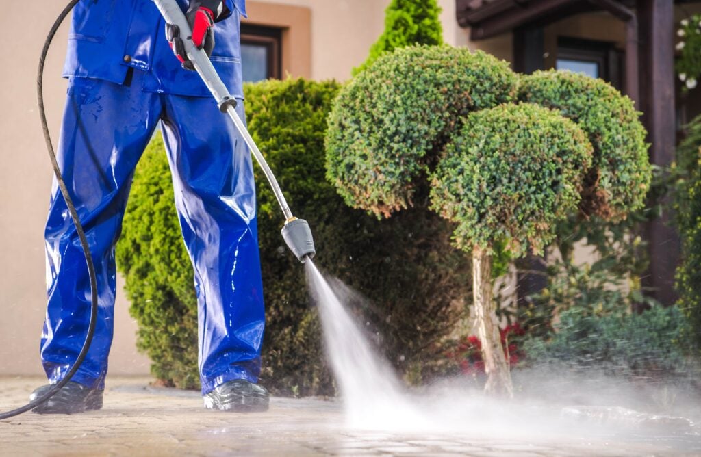 A professional cleaner in blue waterproof gear using a high-pressure washer to clean a stone paver driveway in front of residential landscaping. Oak Run Ocala Exterior Cleaning Services.