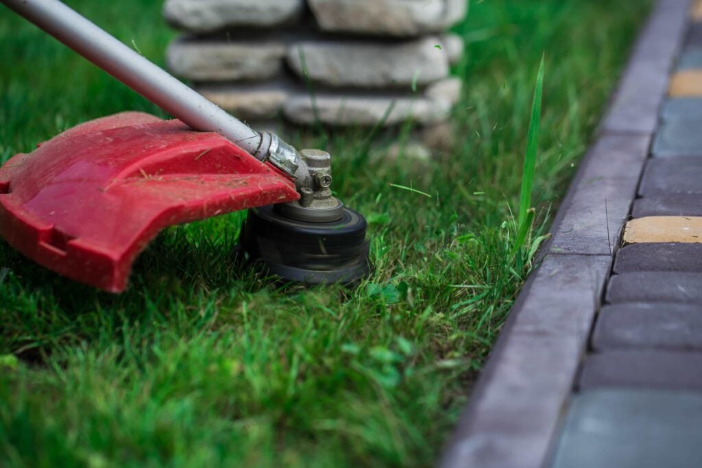 A red string trimmer head cutting grass along a brick walkway and stone garden border, providing professional best lawn service in Stone Creek and edging service in The Villages, Florida.