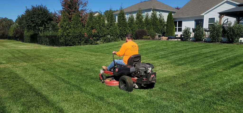 A professional lawn care services technician in Charlotte, NC, wearing an orange shirt while riding a zero-turn commercial mower to create clean stripes on a large, green suburban backyard lawn.